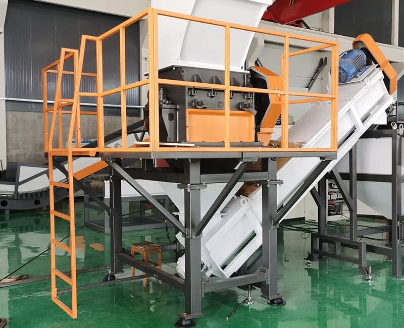 This image features a large industrial shredding or crushing machine. The equipment is elevated on a platform with safety railings and a ladder, providing secure operator access. A conveyor belt extends down from the machine, suggesting it's used to transport processed materials. The hopper at the top is designed to intake large quantities of material. The "Rumtoo" logo and website indicate this machine is part of the company's product line, likely focused on plastic waste management and recycling. This setup looks suitable for processing bulky waste or recyclable materials like plastics or paper, reducing their size for further recycling or disposal.