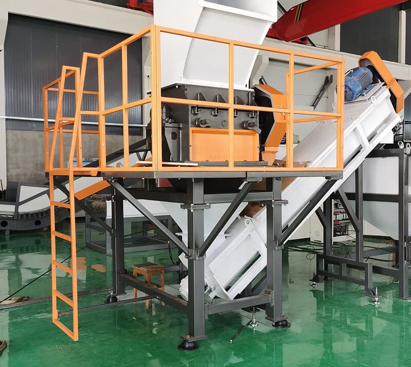 This image features a large industrial shredding or crushing machine. The equipment is elevated on a platform with safety railings and a ladder, providing secure operator access. A conveyor belt extends down from the machine, suggesting it's used to transport processed materials. The hopper at the top is designed to intake large quantities of material. The "Rumtoo" logo and website indicate this machine is part of the company's product line, likely focused on plastic waste management and recycling. This setup looks suitable for processing bulky waste or recyclable materials like plastics or paper, reducing their size for further recycling or disposal.