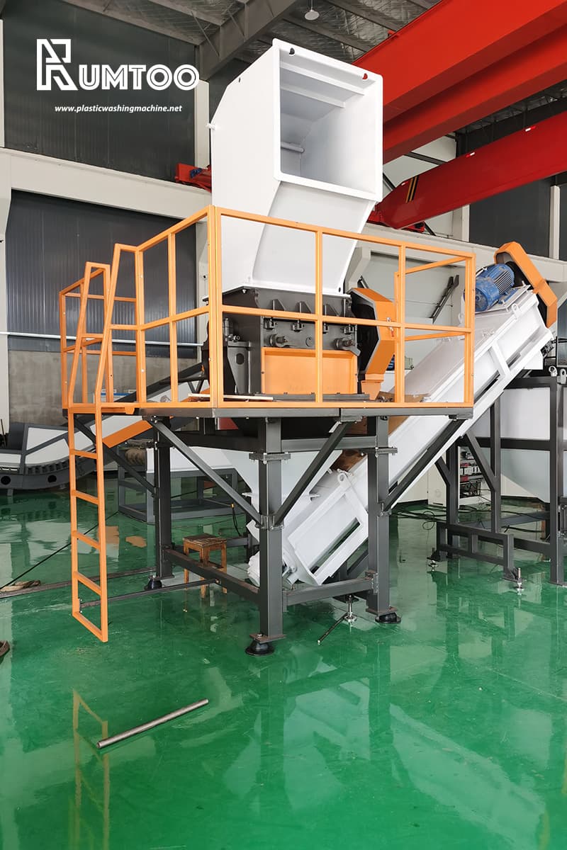 Esta imagem apresenta uma grande máquina industrial de trituração ou esmagamento. O equipamento está elevado numa plataforma com grades de segurança e uma escada, proporcionando um acesso seguro ao operador. Uma correia transportadora estende-se para baixo a partir da máquina, sugerindo que é utilizada para transportar materiais processados. A tremonha no topo foi concebida para receber grandes quantidades de material. O logótipo "Rumtoo" e o sítio Web indicam que esta máquina faz parte da linha de produtos da empresa' provavelmente centrada na gestão e reciclagem de resíduos de plástico. Esta configuração parece adequada para processar resíduos volumosos ou materiais recicláveis, como plásticos ou papel, reduzindo o seu tamanho para posterior reciclagem ou eliminação.