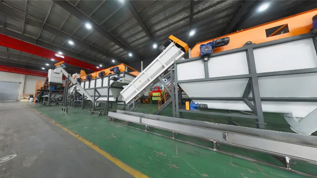 een industrieel plasticrecyclingwassysteem of recyclinginstallatie. De grote machines met transportbanden en sorteerelementen zijn kenmerkend voor apparatuur die wordt gebruikt om plasticafvalmaterialen te wassen, sorteren en verwerken als onderdeel van het plasticrecyclingproces. Deze systemen zijn ontworpen om grote volumes plastic artikelen of balen efficiënt te verwerken, ze op type te scheiden, verontreinigingen te verwijderen via was-/spoelfasen, en de gereinigde plasticmaterialen voor te bereiden voor verdere recycling en verwerking tot nieuwe plasticproducten of grondstoffen. De oranje en grijze kleurstelling van de machines is ook kenmerkend voor veel plasticrecyclingfaciliteiten en -systemen.